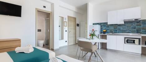 A modern hotel room with two beds, a dining area, and a kitchenette.