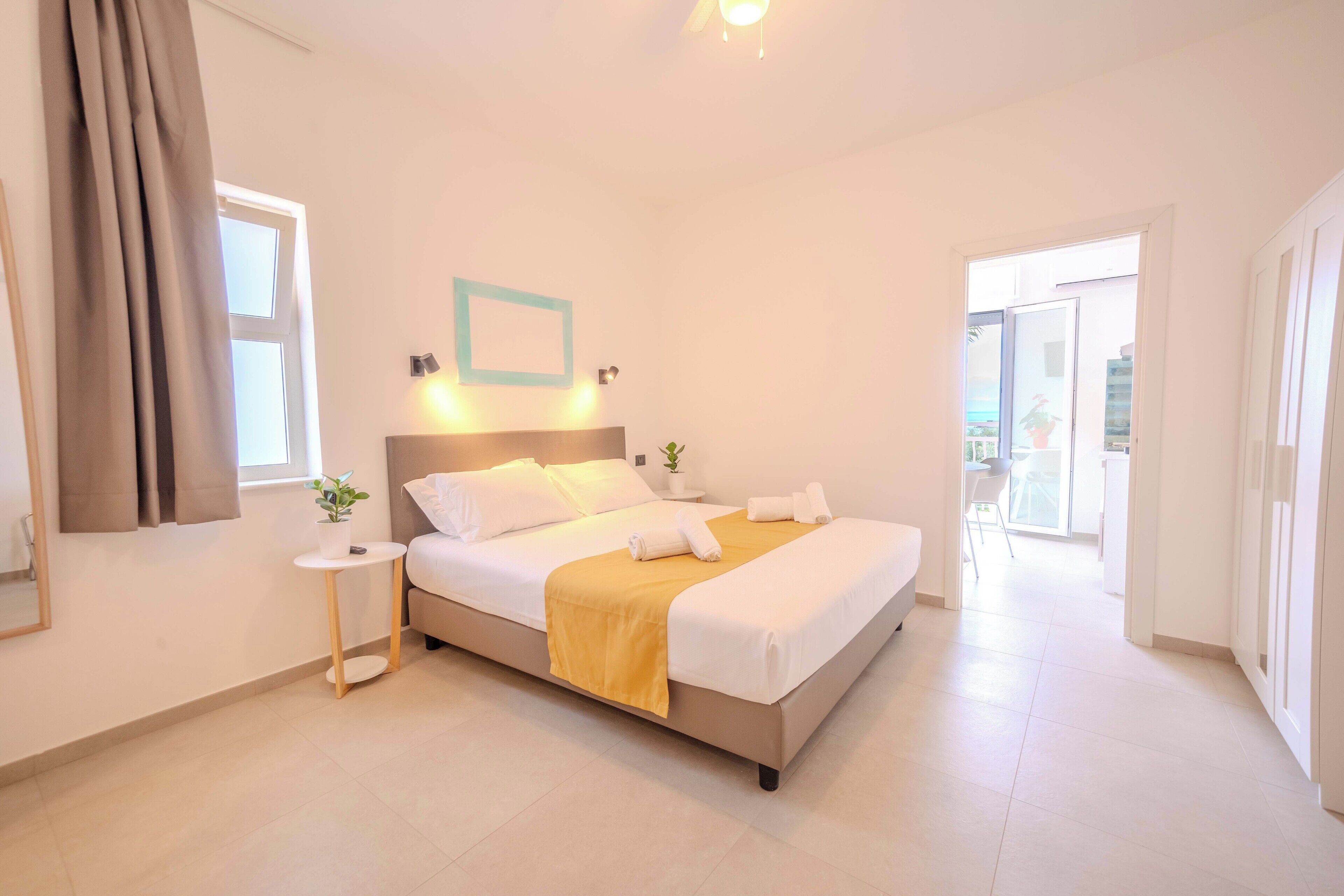 A modern hotel room with a large bed, a bedside table with a plant, a small table, and a view of the outside balcony.