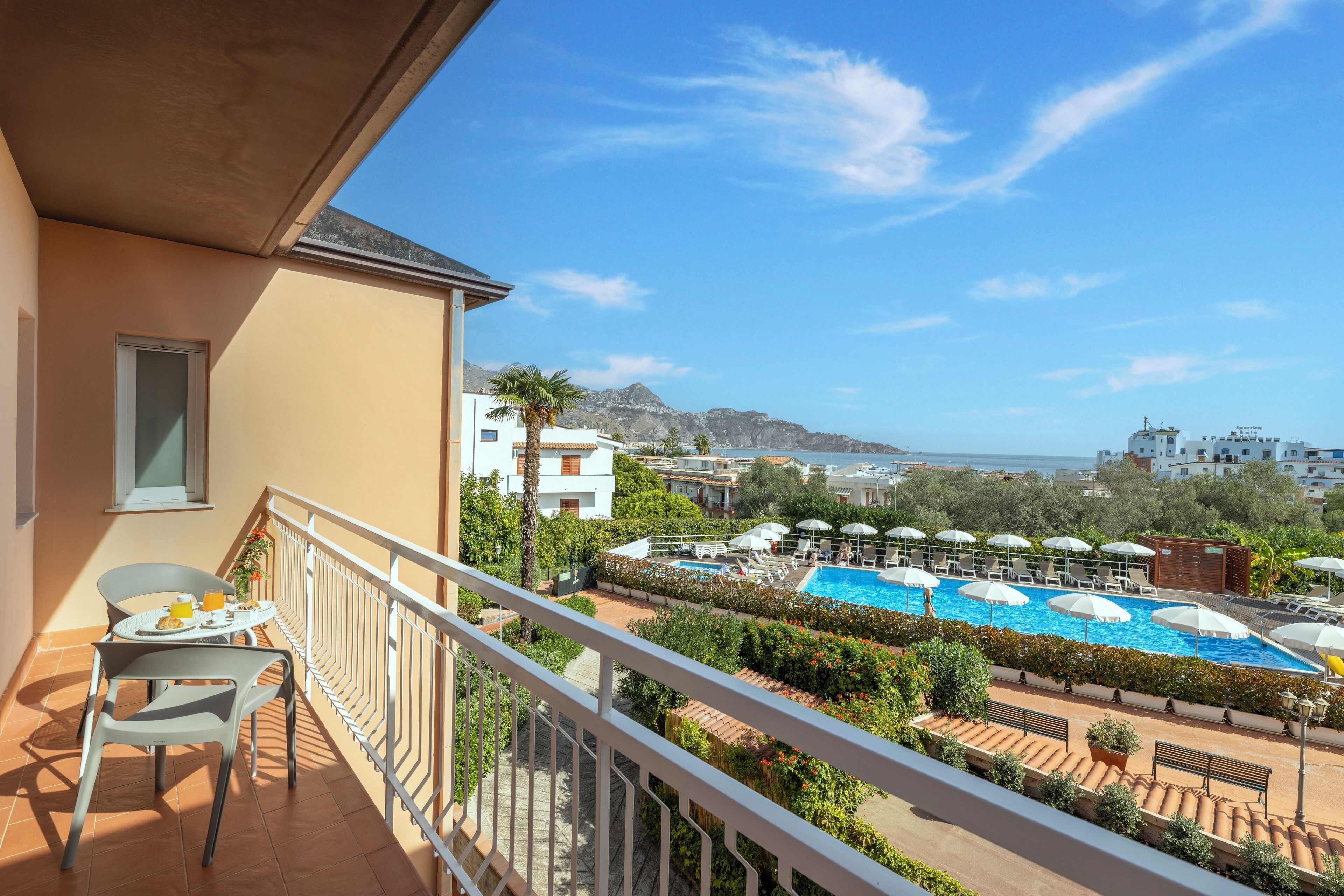 Un balcón con vistas a una piscina, mobiliario exterior y el paisaje de montañas al fondo.