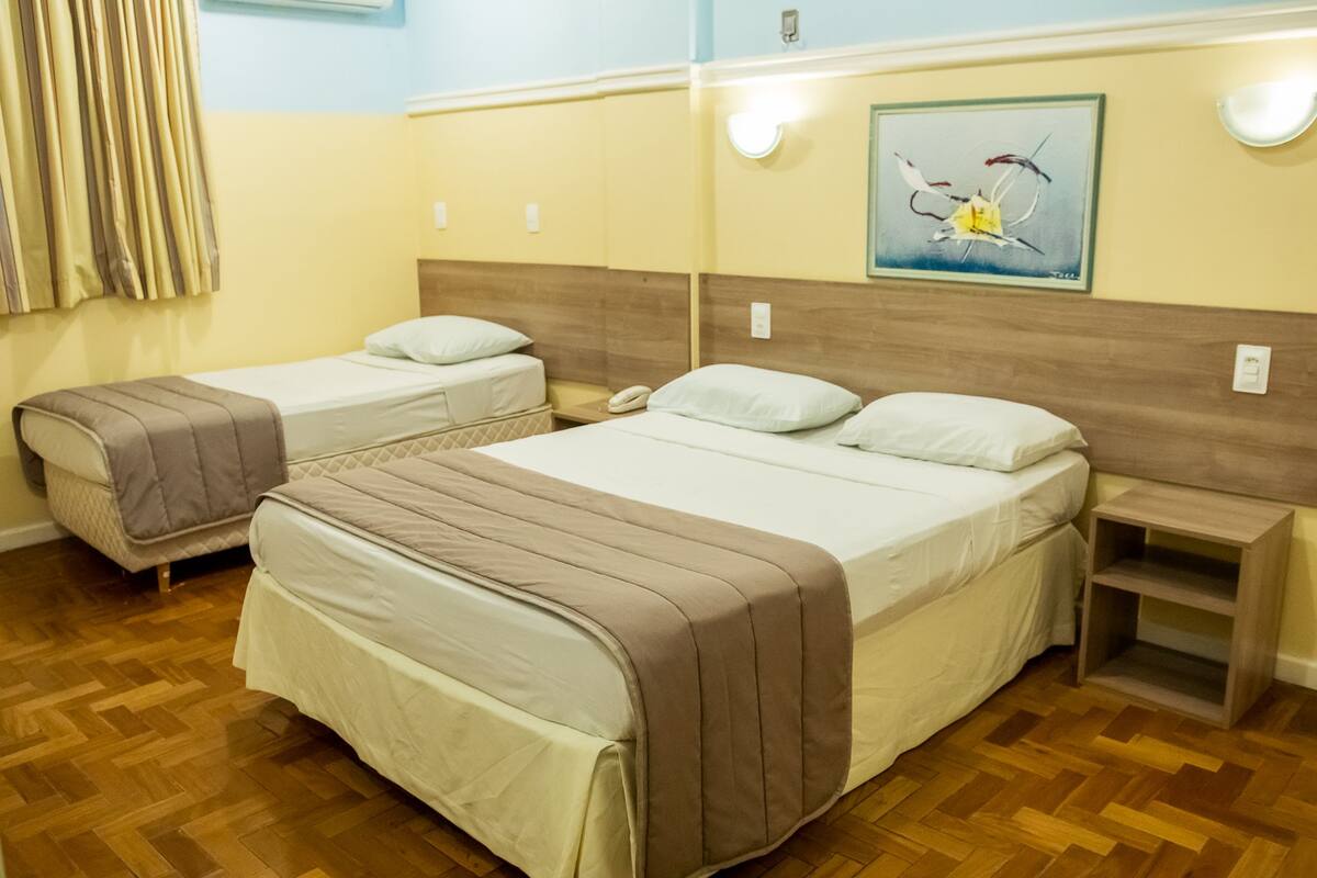 A hotel room with two single beds, wooden headboards, a small bedside table with a lamp, and a painting on the wall.