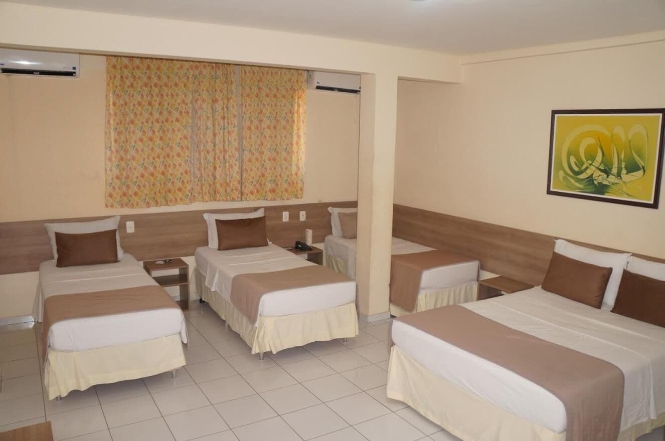 A hotel room with four single beds, a large window with patterned curtains, and a framed abstract painting on the wall.