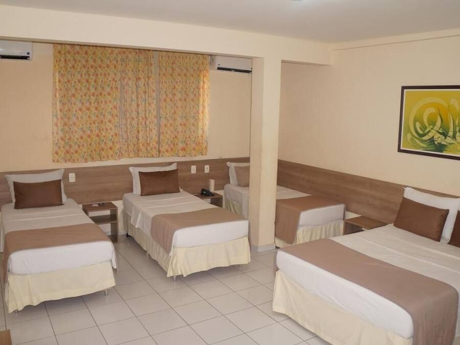 A hotel room with four single beds, a large window with patterned curtains, and a framed abstract painting on the wall.
