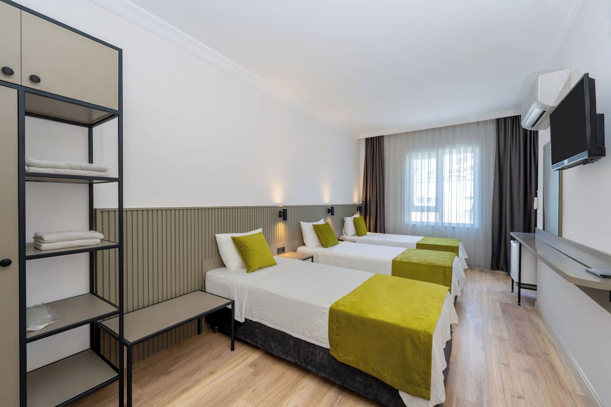 A hotel room with three beds, a TV, a shelf with towels, and a window with curtains.