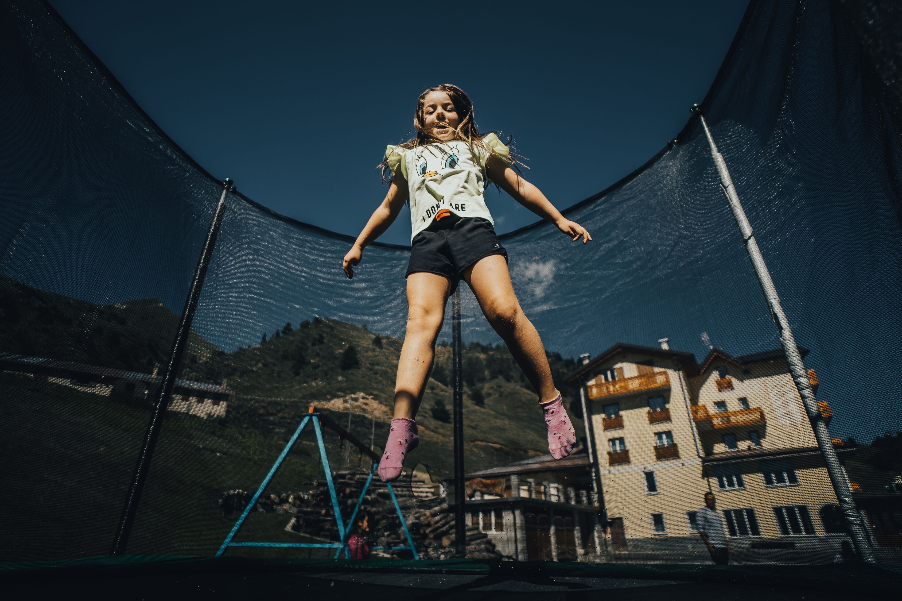A person jumping on a trampoline with a netted enclosure, set against a backdrop of buildings and hills.