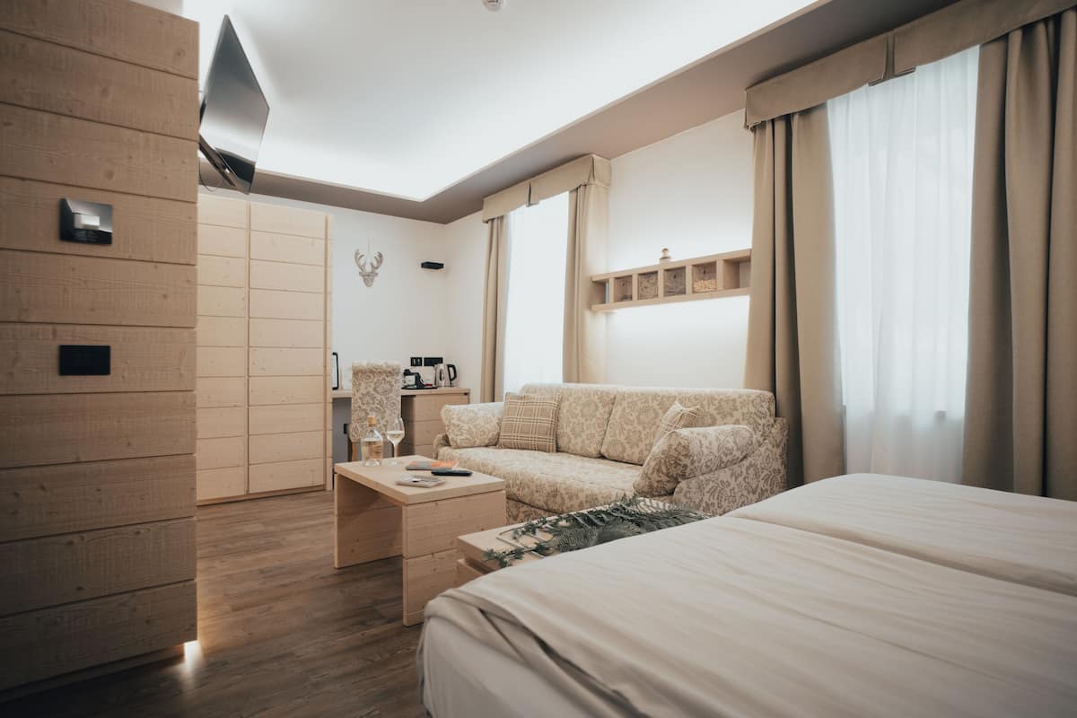 A modern hotel room with a wooden wardrobe, a sofa, a bed, and a small table with a bottle and glasses.