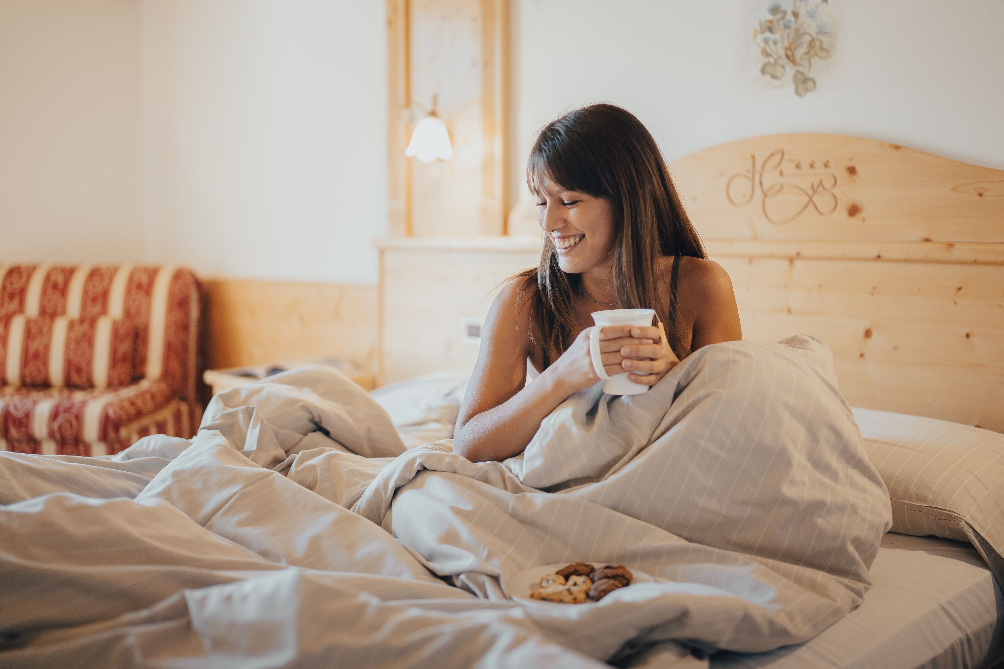 Une personne est assise sur un lit, une tasse à la main, dans une pièce dont le lit est doté d’une tête de lit en bois et dont un mur est décoré.