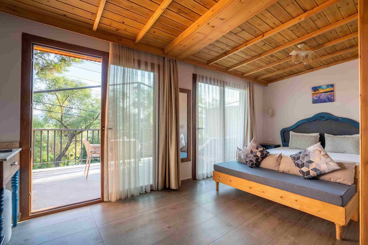 Ein Schlafzimmer mit Holzbalkendecke, einem Bett mit gemusterten Kissen, einer Bank und Blick ins Freie durch große Fenster.