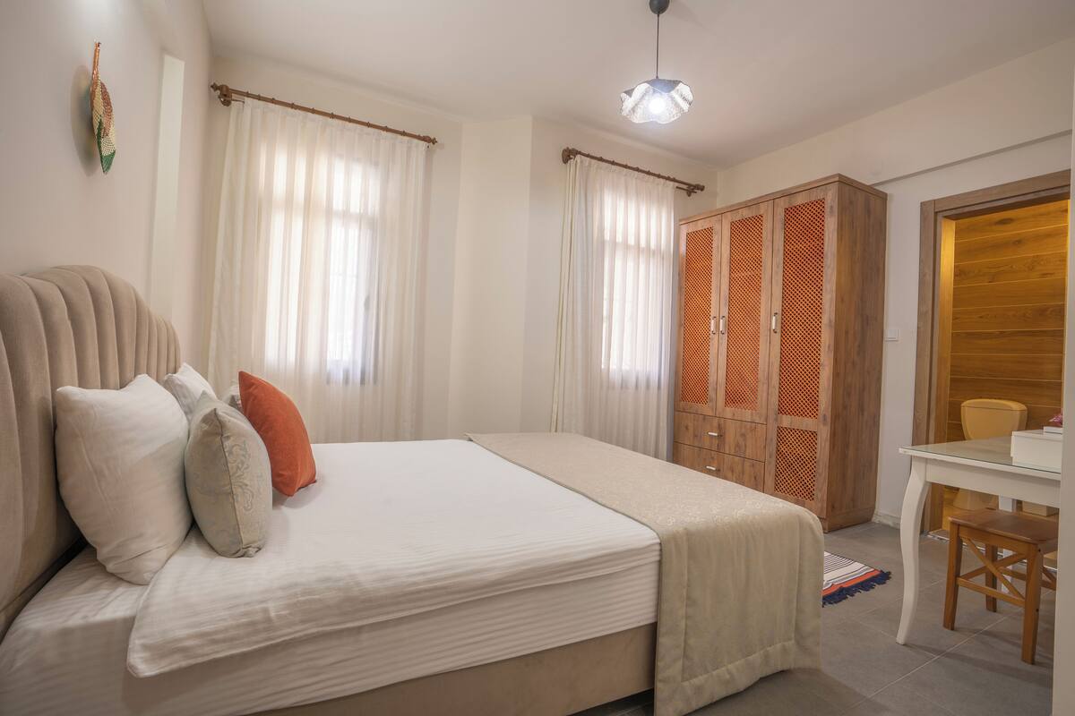 Ein Schlafzimmer mit Bett, Kissen, Vorhängen, einem Schrank, einem Schreibtisch und einem Sessel.