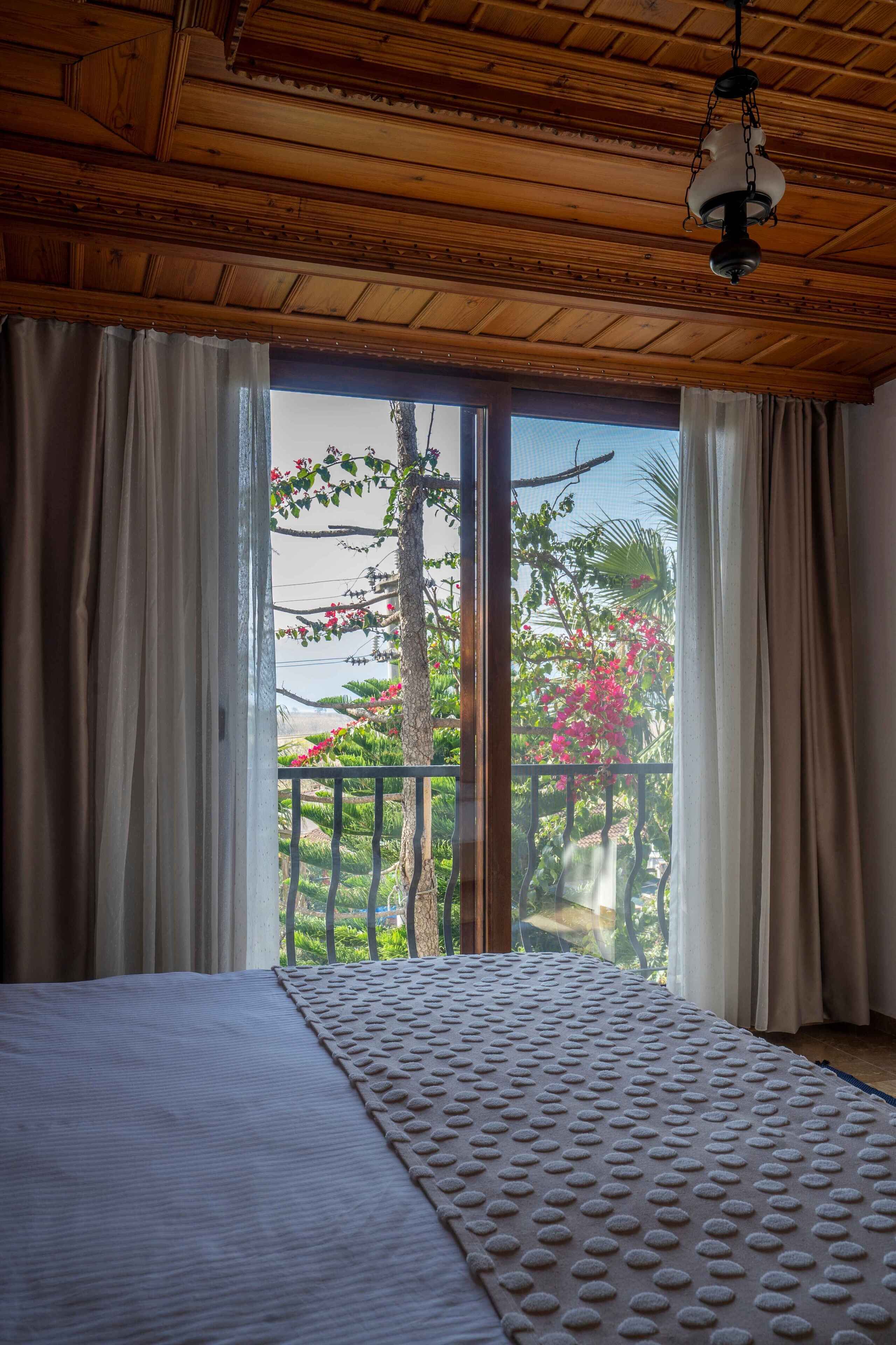 A room with a bed, wooden ceiling, and a view of a balcony with plants and flowers.