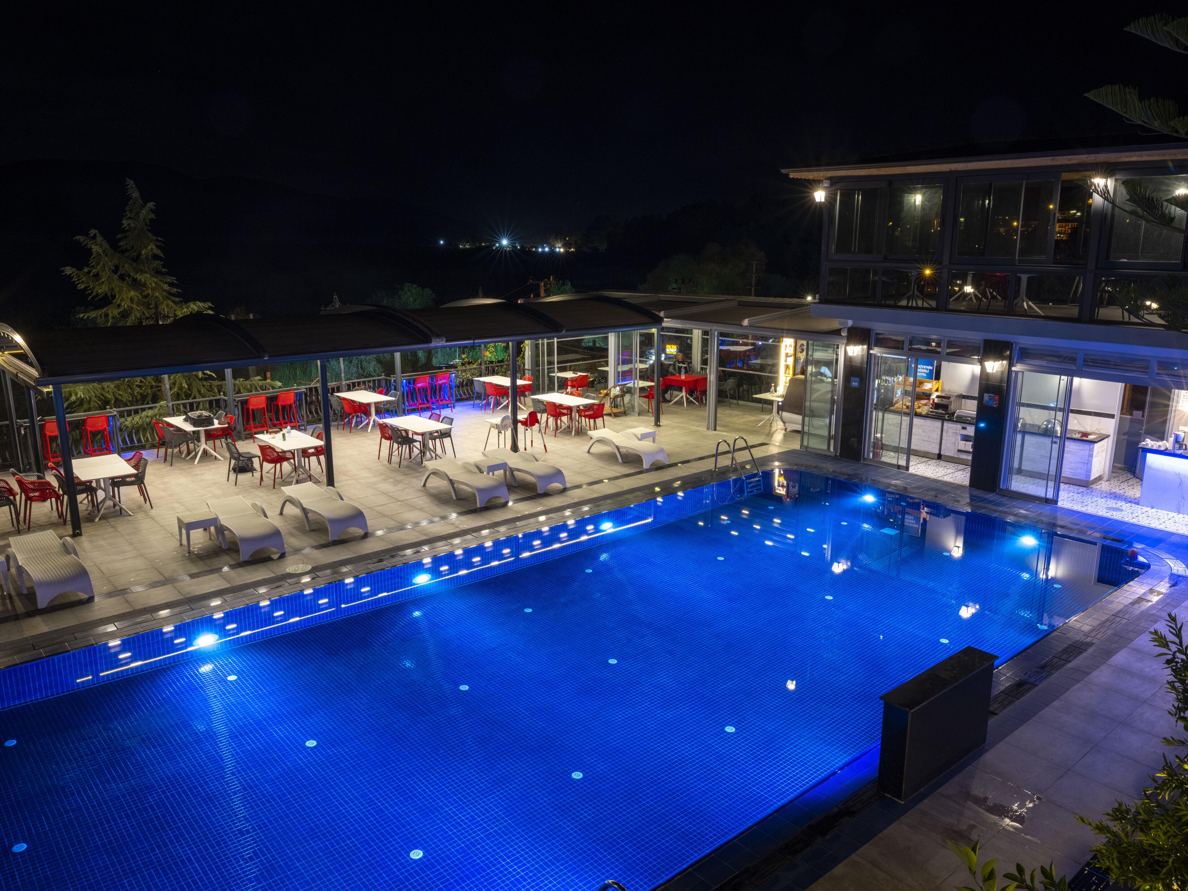 A swimming pool with lounge chairs and tables, surrounded by a covered patio area with red chairs and a restaurant in the background.