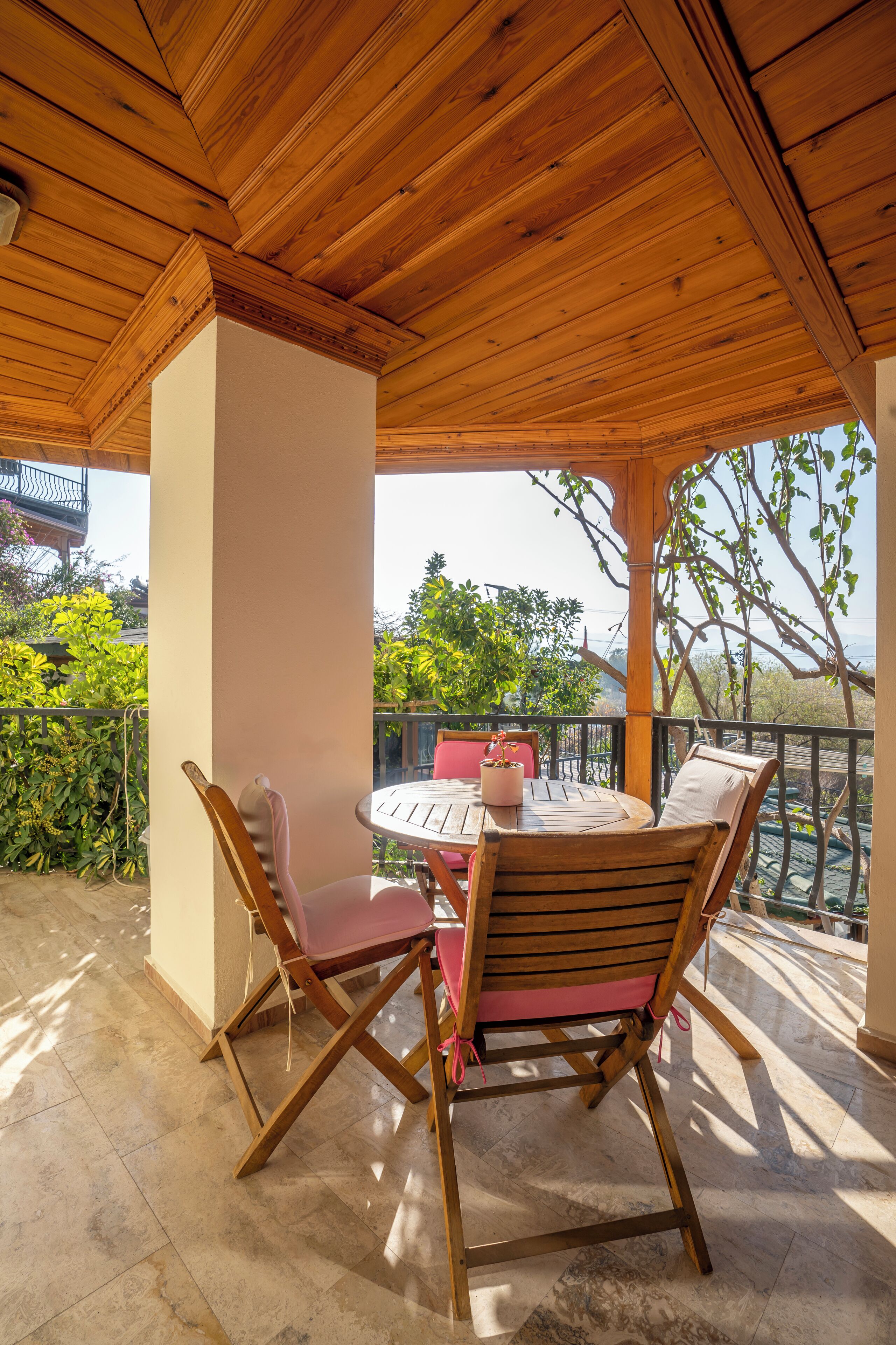 A covered patio with a wooden table and chairs, a view of greenery, and a balcony railing.