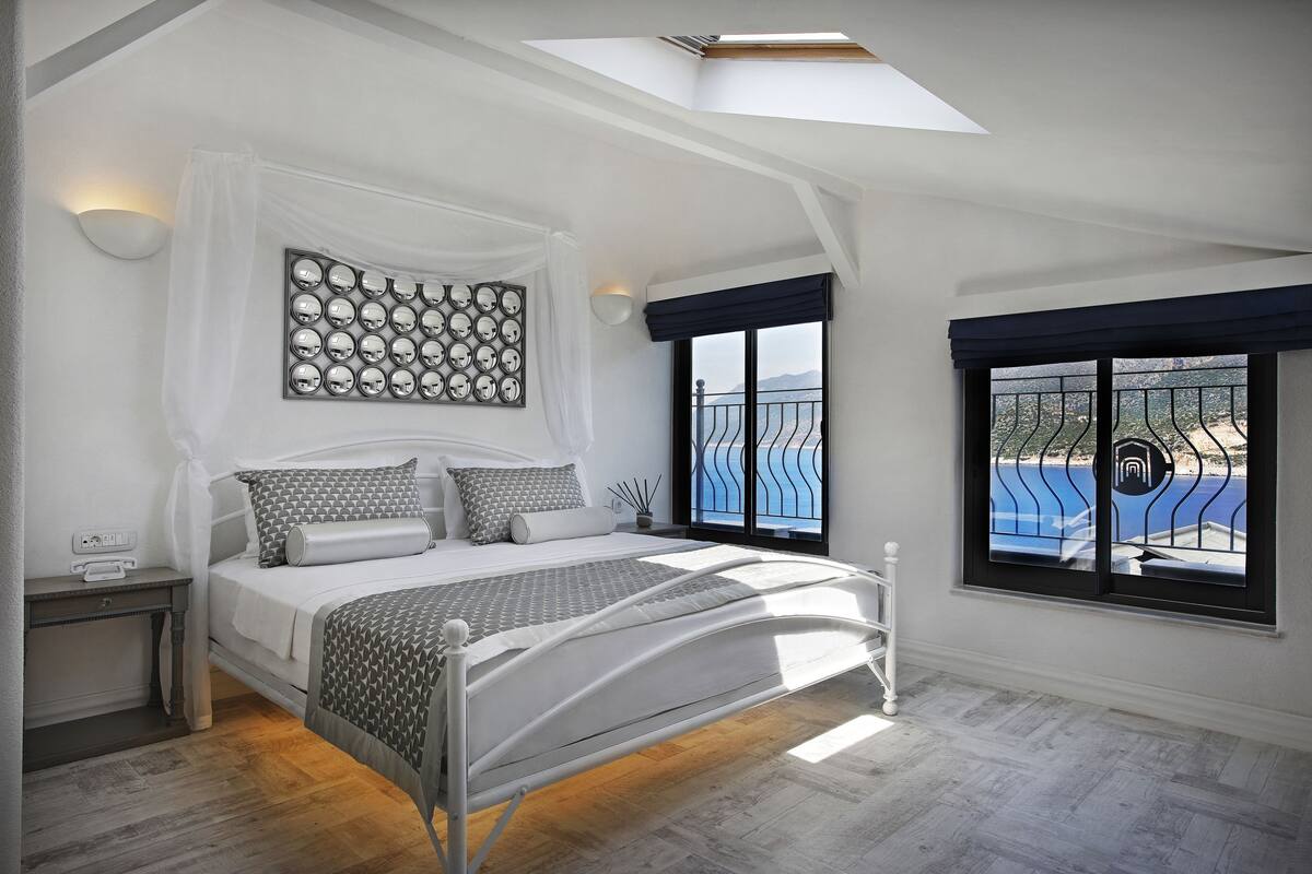 A bedroom with a bed, bedside table, and a view of the sea through a window.