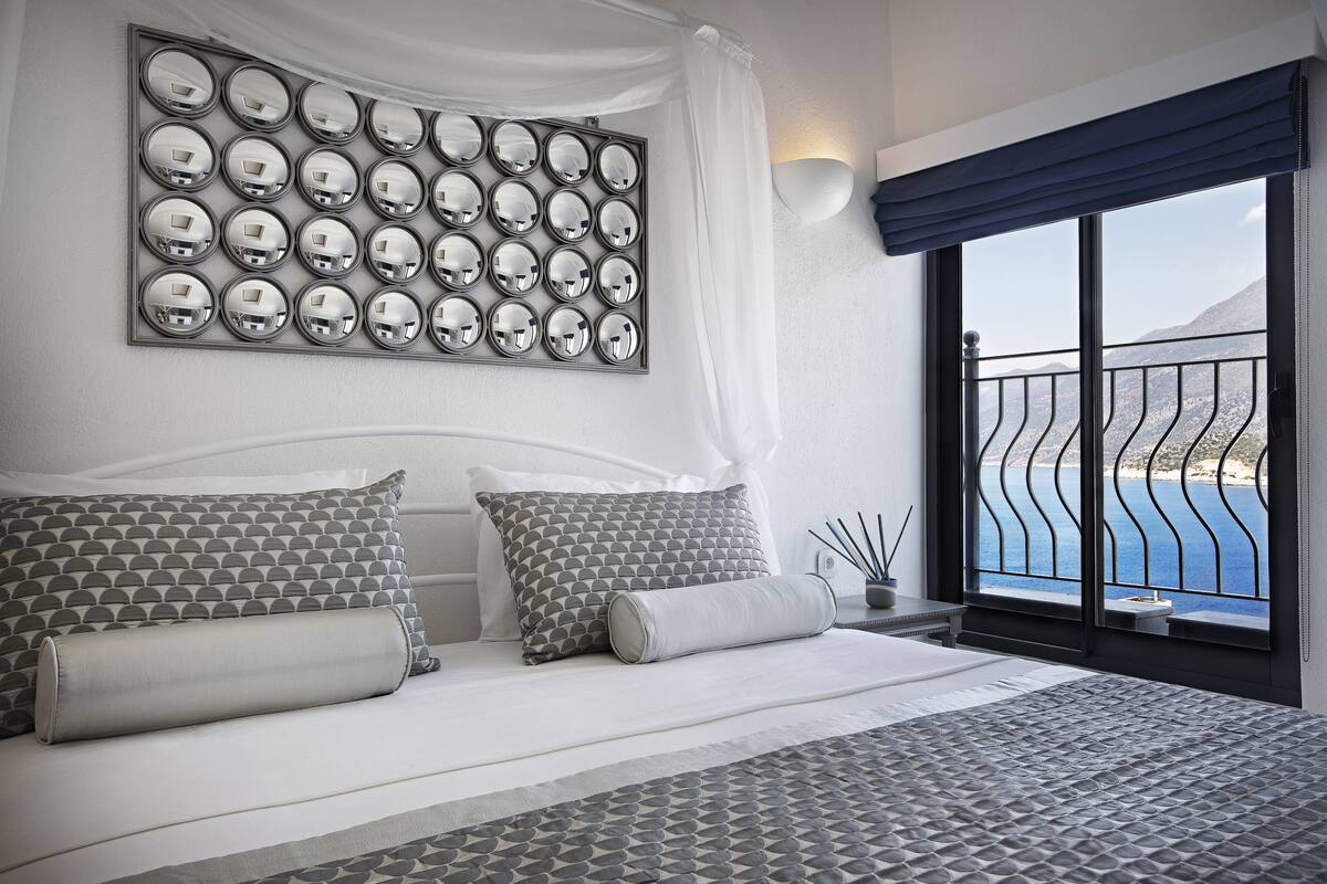A bedroom with a large bed, a wall-mounted mirror, a view of the sea, and a balcony with a railing.
