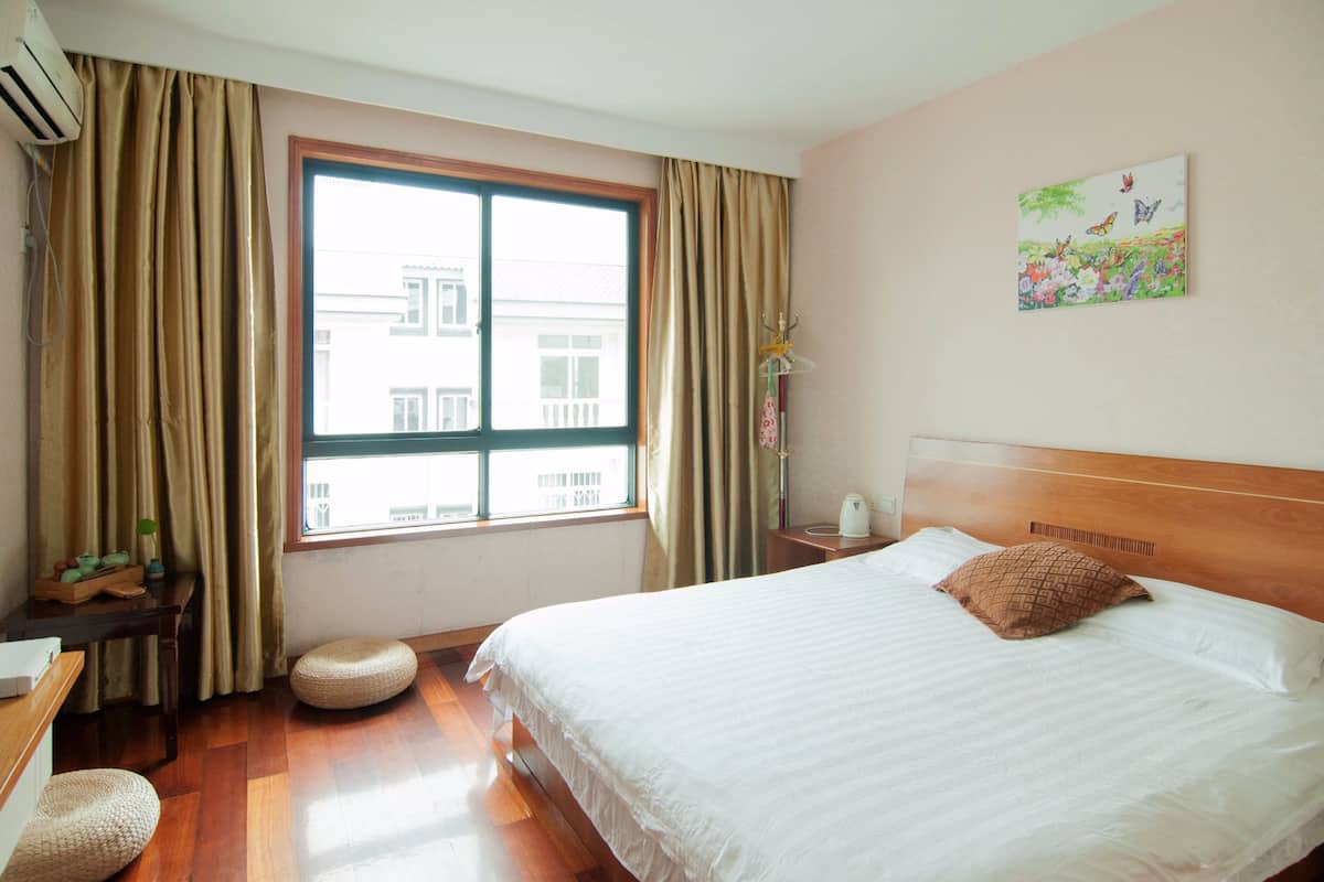 A hotel room with a large window, a bed with white bedding, a wooden headboard, a bedside table with a lamp, and a painting on the wall.