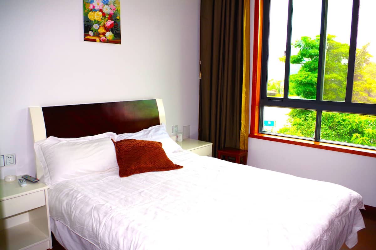 A neatly made bed with white linens and a red accent pillow, a bedside table with a remote control, and a window with a view of greenery.
