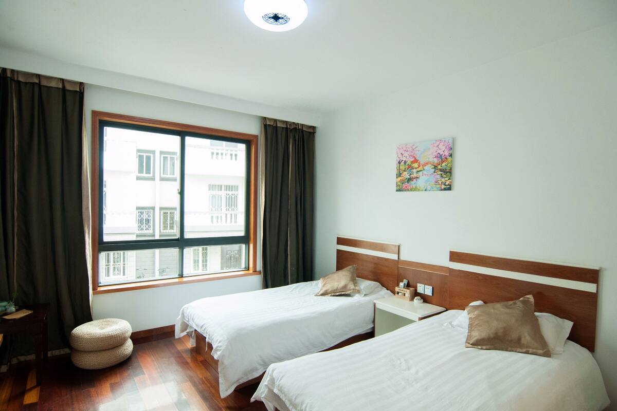 A hotel room with two beds, a window with curtains, a small round ottoman, and a painting on the wall.