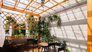 Ein überdachter Patio-Bereich mit Holzboden, zwei schwarzen Stühlen, einem kleinen Tisch und einer Holz-Rankgitter mit Kletterpflanzen.