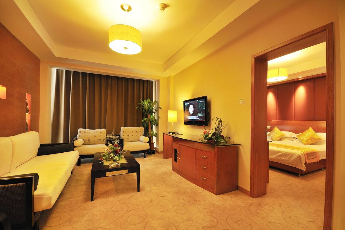 Habitación de hotel con sofá, sillón, mesa de centro, televisor y cama con cabecero.