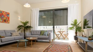 Uma sala de estar com um sofá cinzento, uma mesa de centro redonda e um tapete estampado. Há duas plantas em vasos, uma mesa de madeira com cadeiras e uma janela grande com persianas.