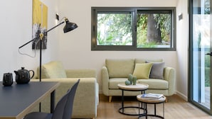 Uma sala de estar acolhedora com um sofá, uma mesa de centro e uma escrivaninha com uma luminária. Há uma janela com vista para árvores e uma porta à direita.