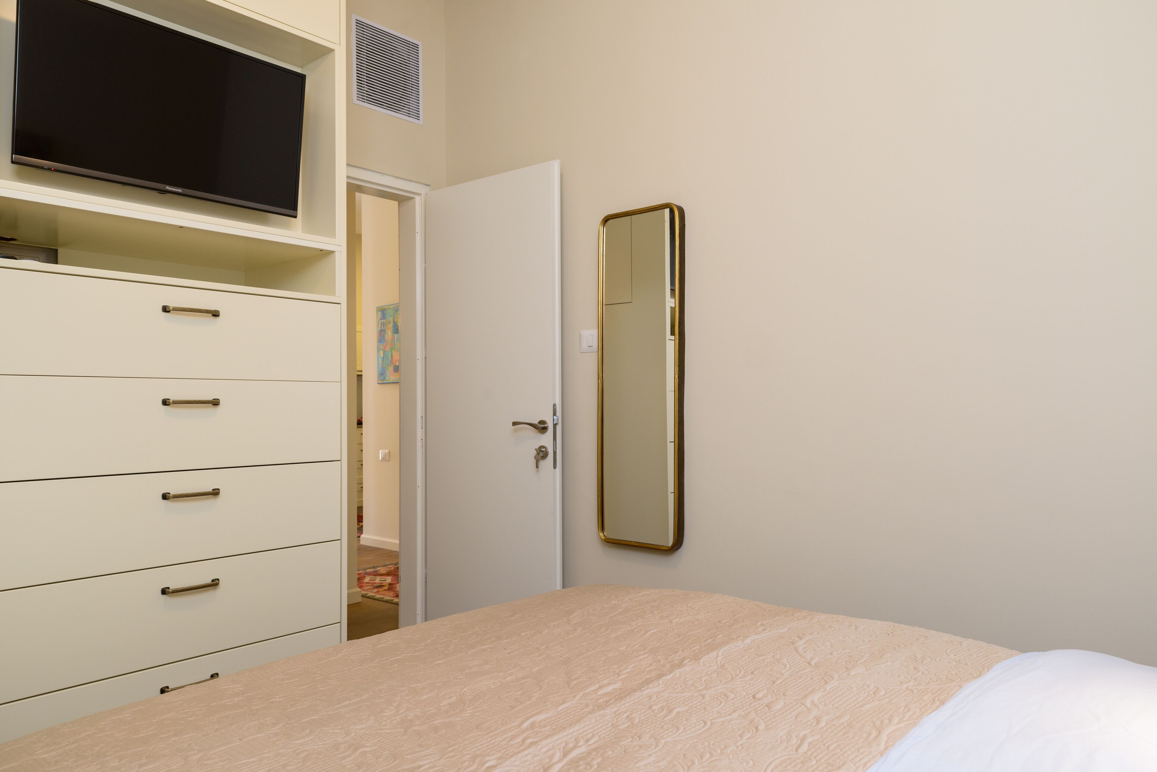 A bedroom with a bed, a dresser with drawers, a television on a shelf, and a door with a mirror.