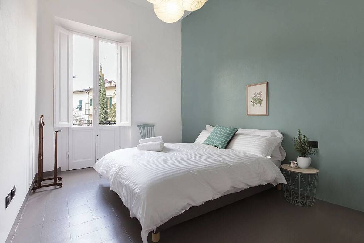 A bedroom with a bed, a window, a door, a bedside table, and a wall-mounted picture.