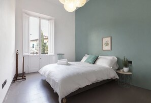 A bedroom with a bed, a window, a door, a bedside table, and a wall-mounted picture.