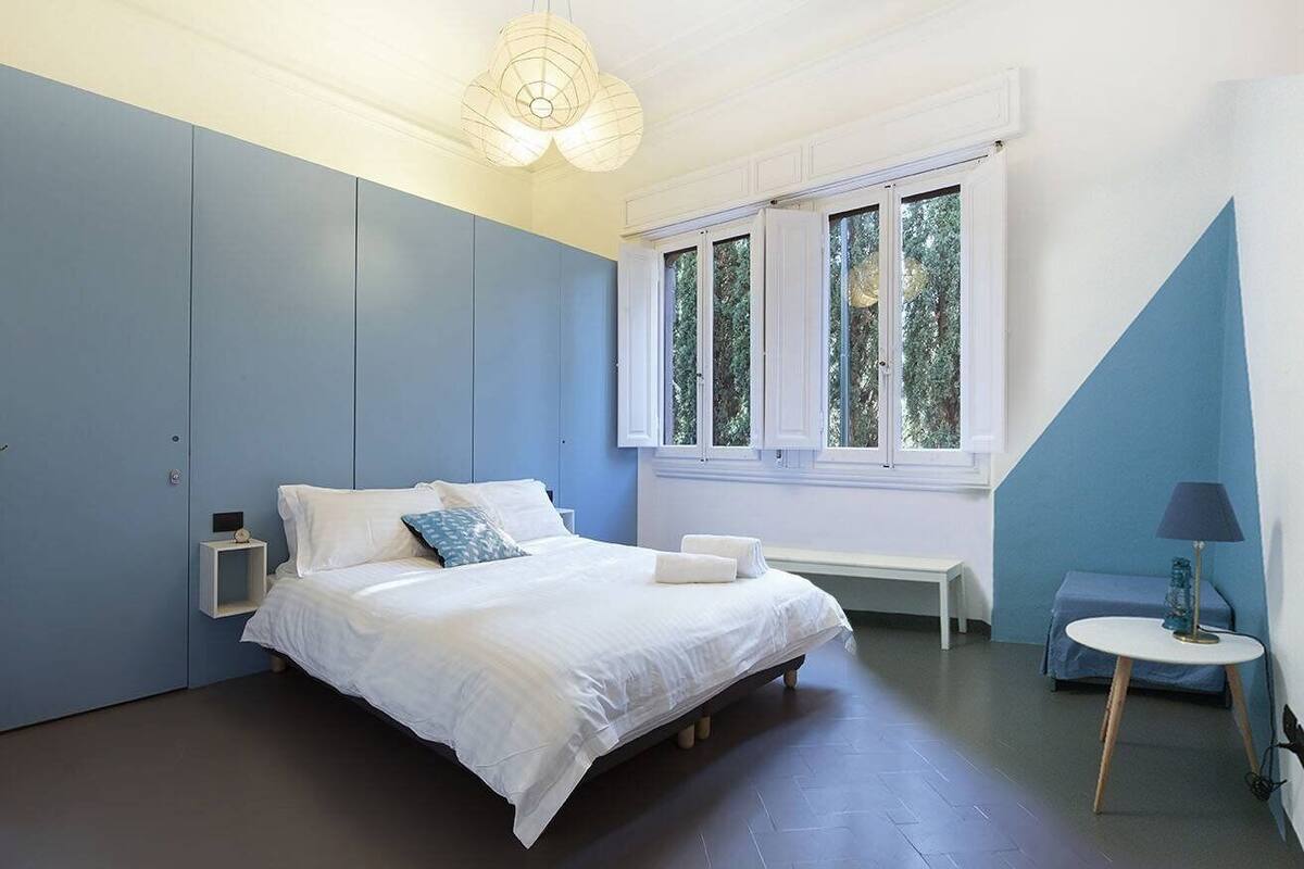 A bedroom with a bed, a bedside table, a lamp, a window, and a bench.