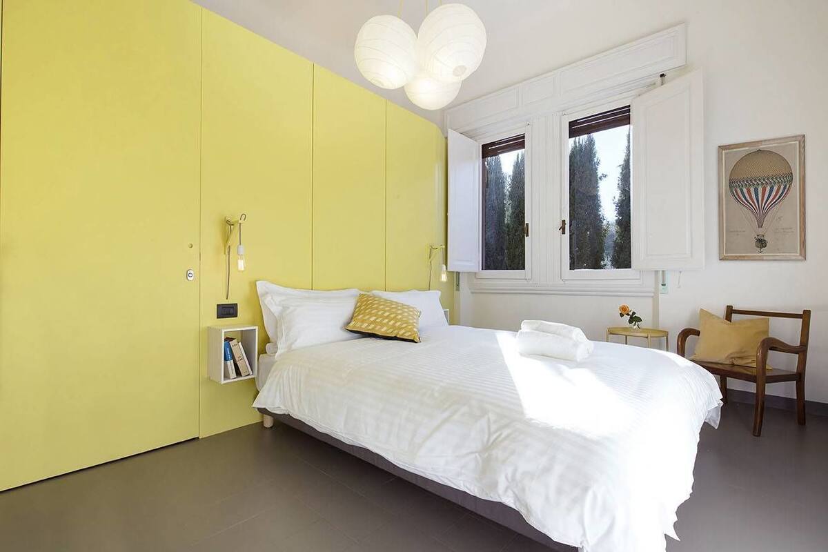 A bedroom with a large bed, a yellow wardrobe, a small bedside table, and a window with a view of trees.