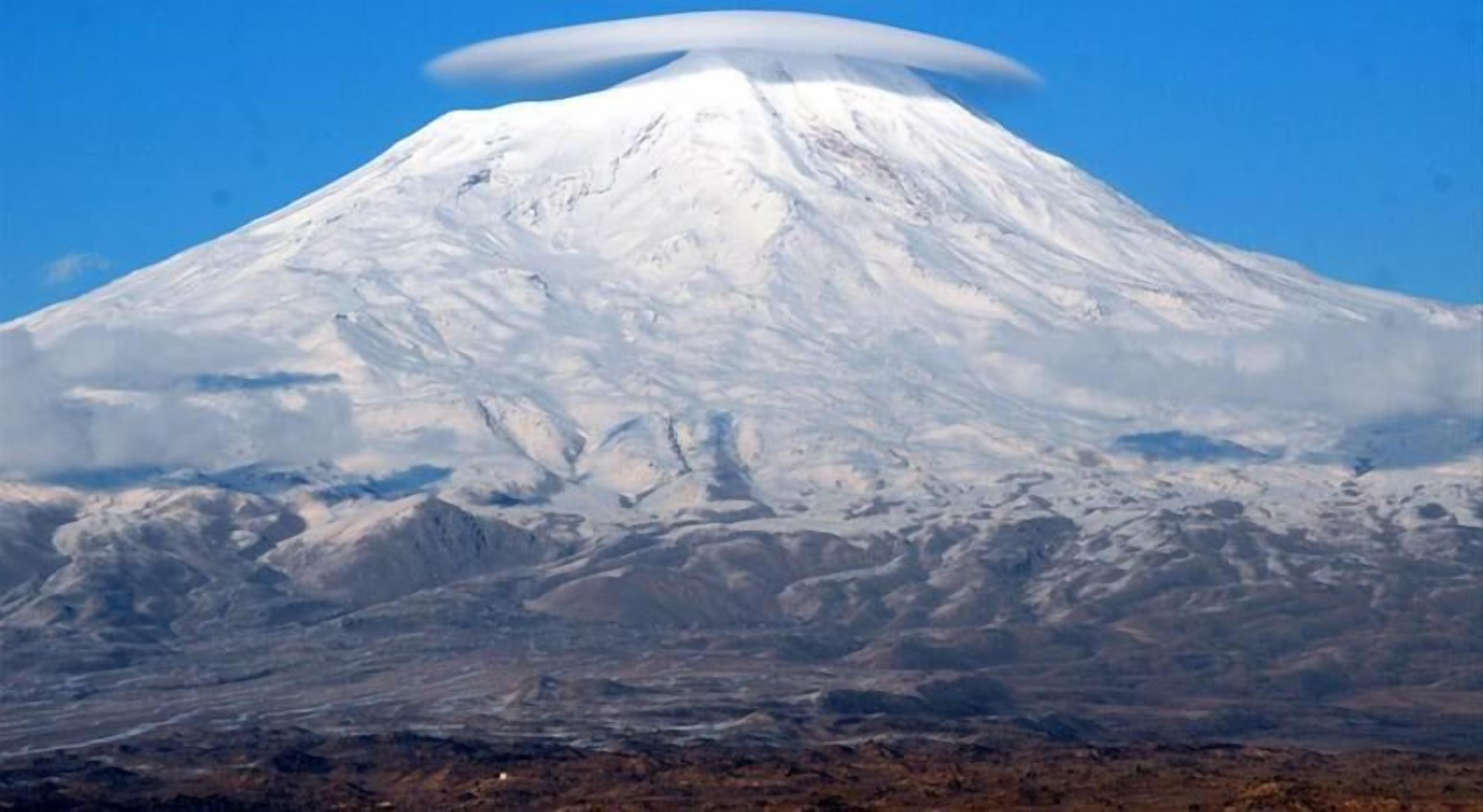 Uma montanha coberta de neve com formato cônico característico e topo plano, rodeada por uma paisagem árida.