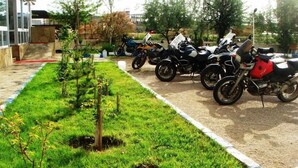 A row of motorcycles parked in a covered area with a grassy path and small trees. - Butik Ertur Hotel (Dogubayazit)