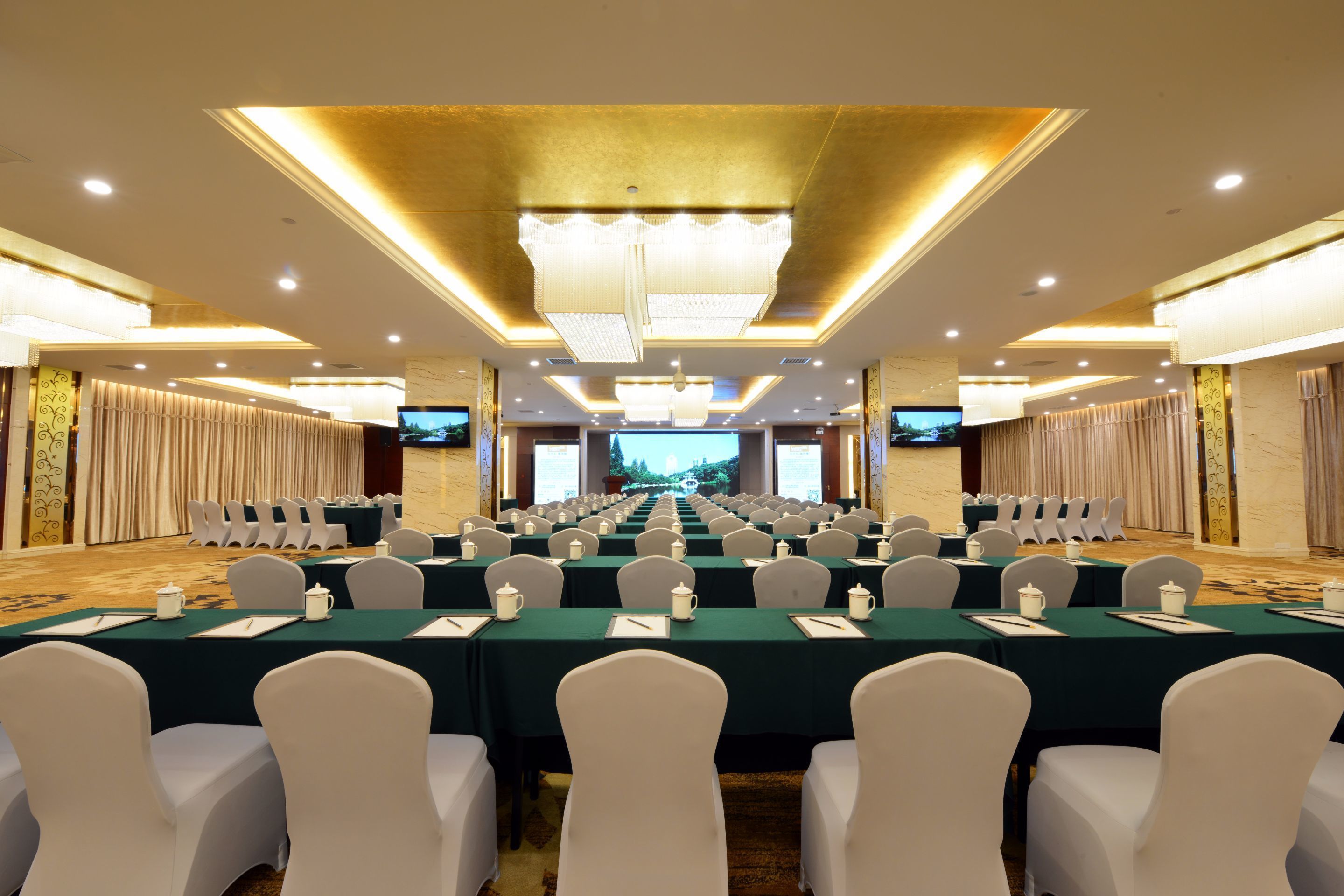 A conference room with rows of tables and chairs, a large screen at the front, and a chandelier.