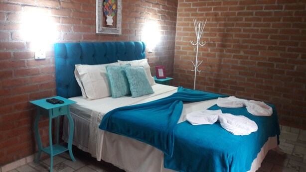 A bedroom with a brick wall, a blue upholstered headboard, a white bedspread, and a teal throw blanket. - Toca Hospedaria (São Bento do Sapucaí)