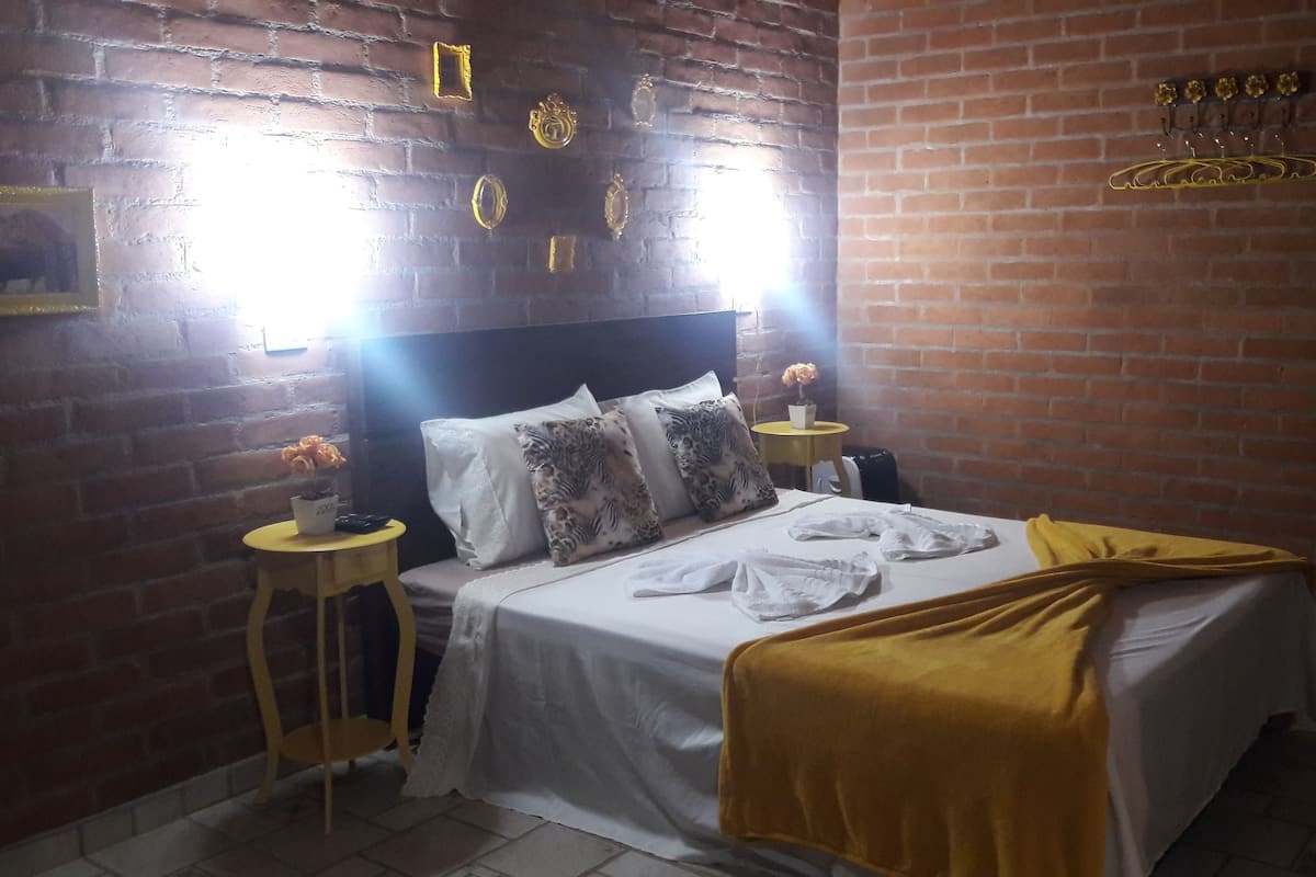 Een kamer met een bakstenen muur, een bed met wit beddengoed, een gele plaid en een klein tafeltje met een bloevas.