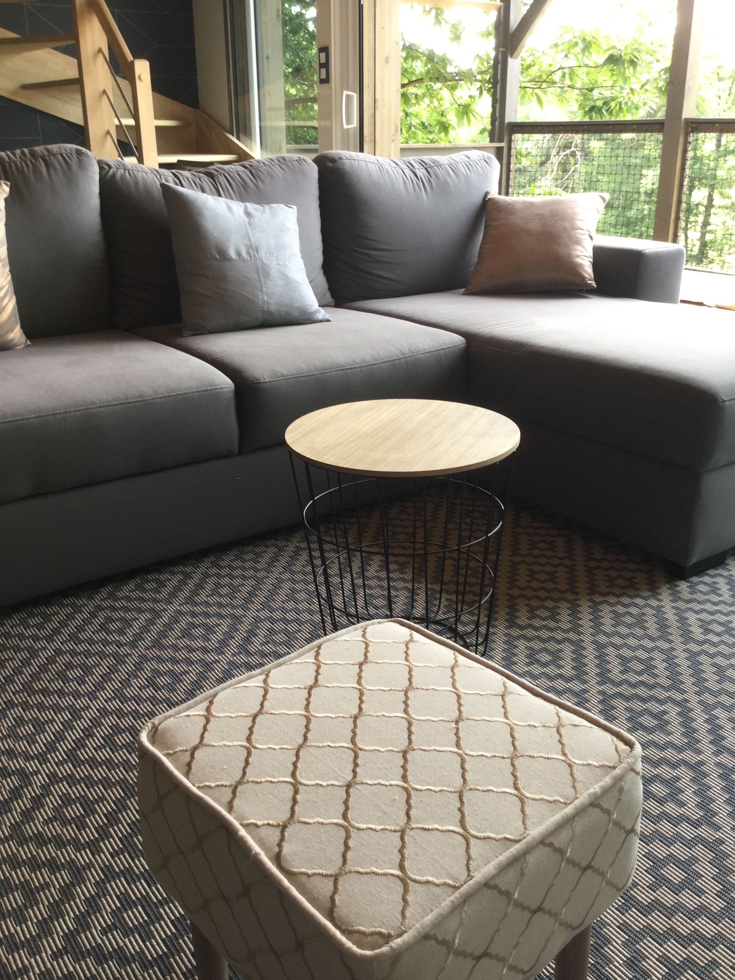 A modern living room with a grey sectional sofa, a round wooden coffee table, and a patterned ottoman.