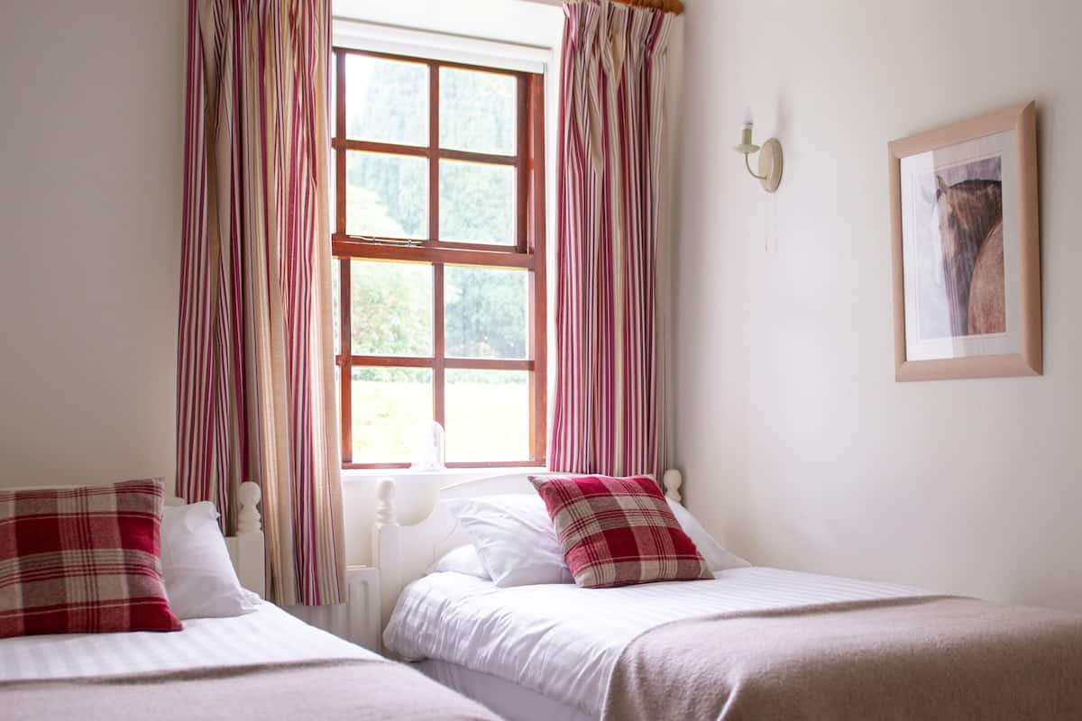 A room with two beds, a window with curtains, a wall-mounted light, and a framed picture.