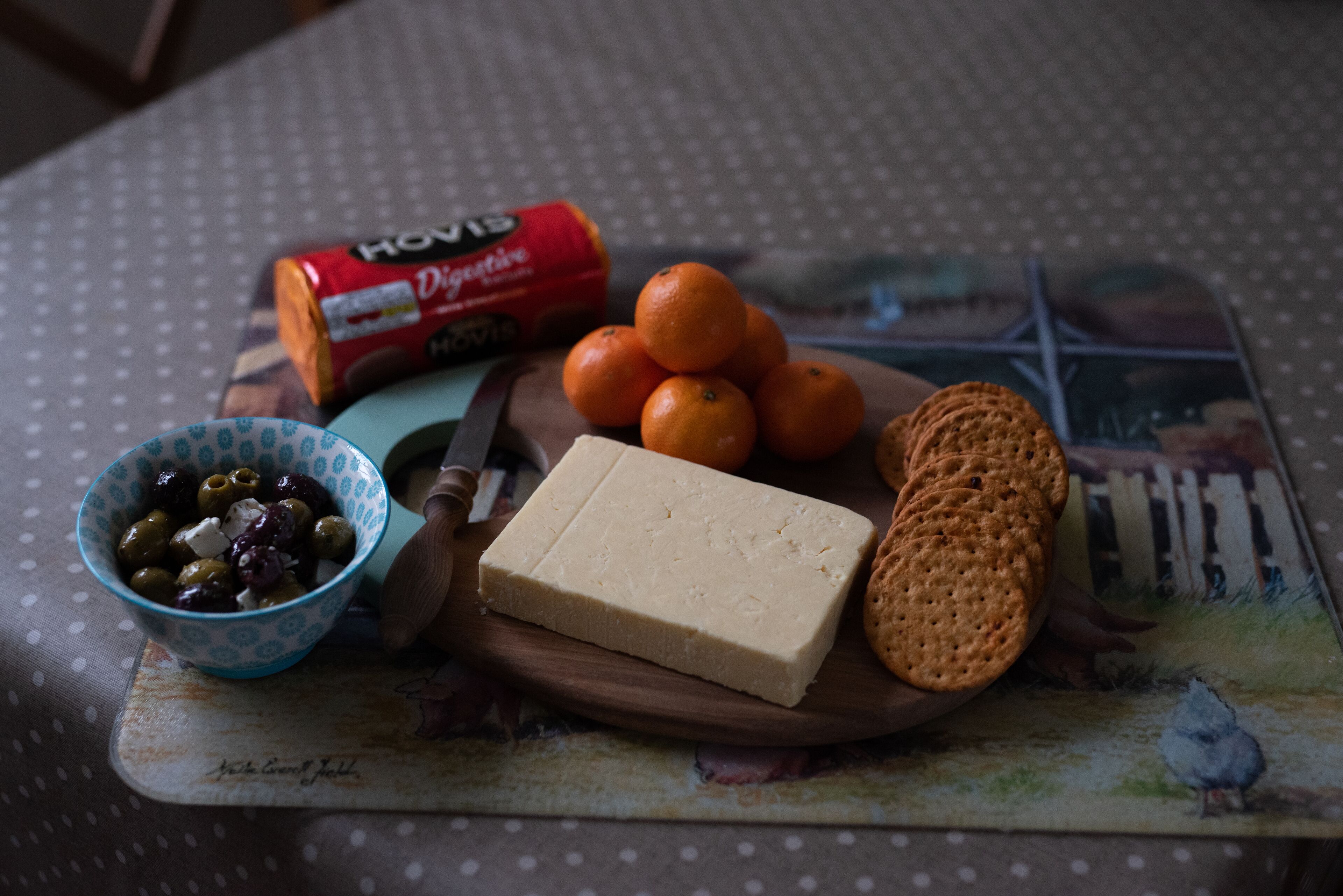 Una tabla de madera con queso, galletas y naranjas sobre una mesa con mantel de lunares.