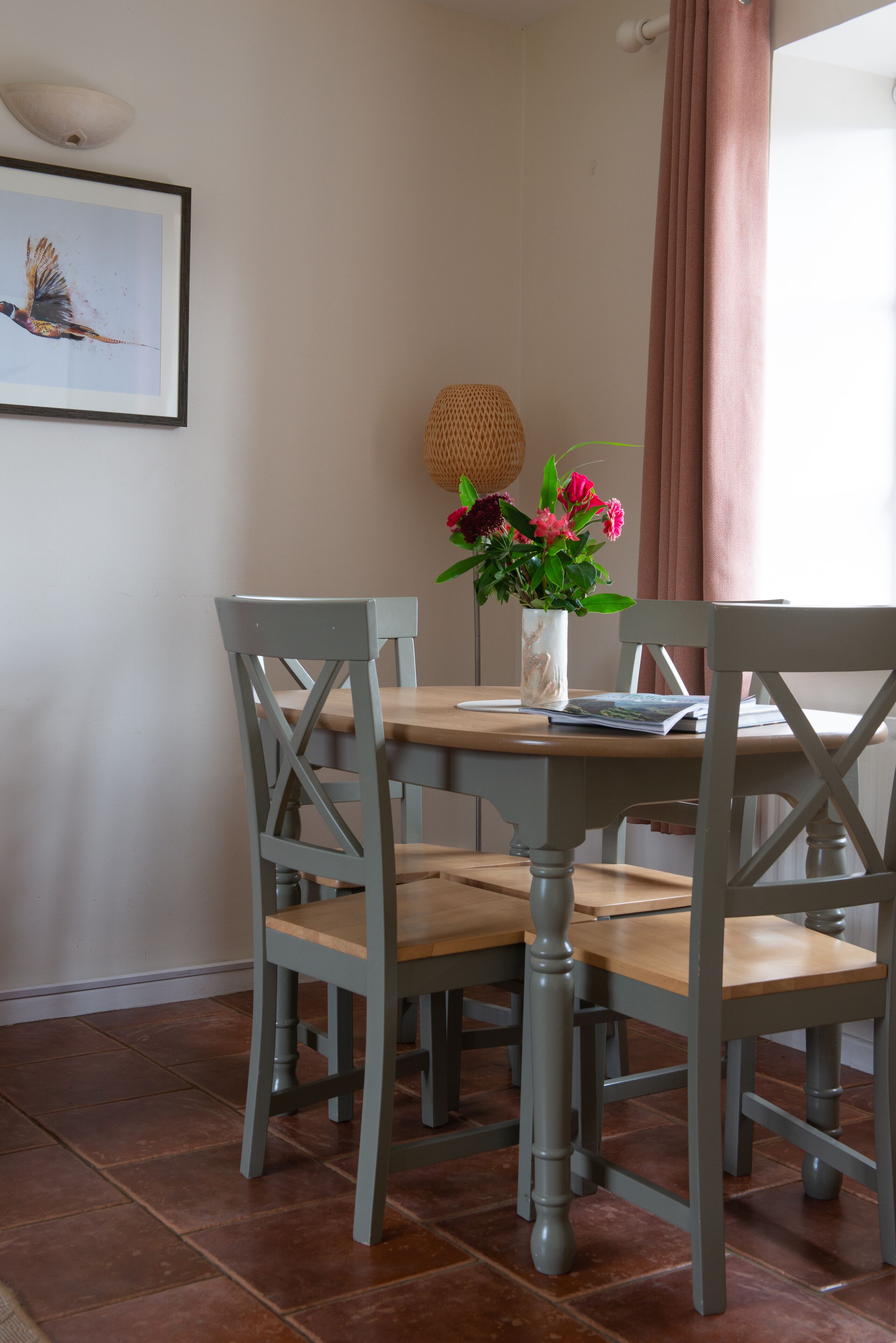 A dining area with a wooden table, four chairs, a vase with flowers, and a framed picture on the wall.