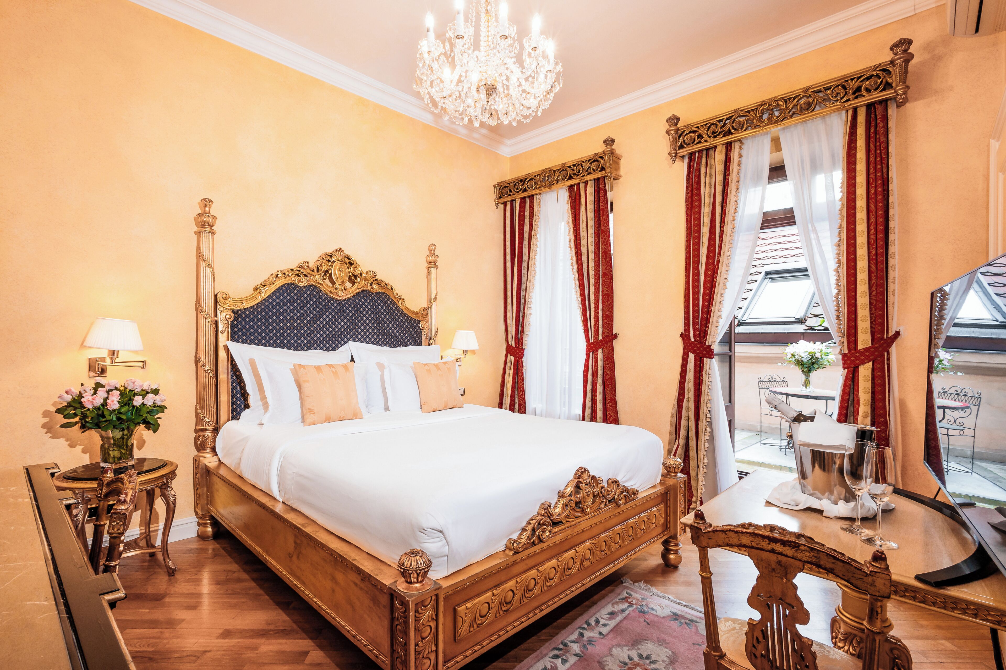 A hotel room with a large bed, a chandelier, a dining table with chairs, and a view of the outdoors.