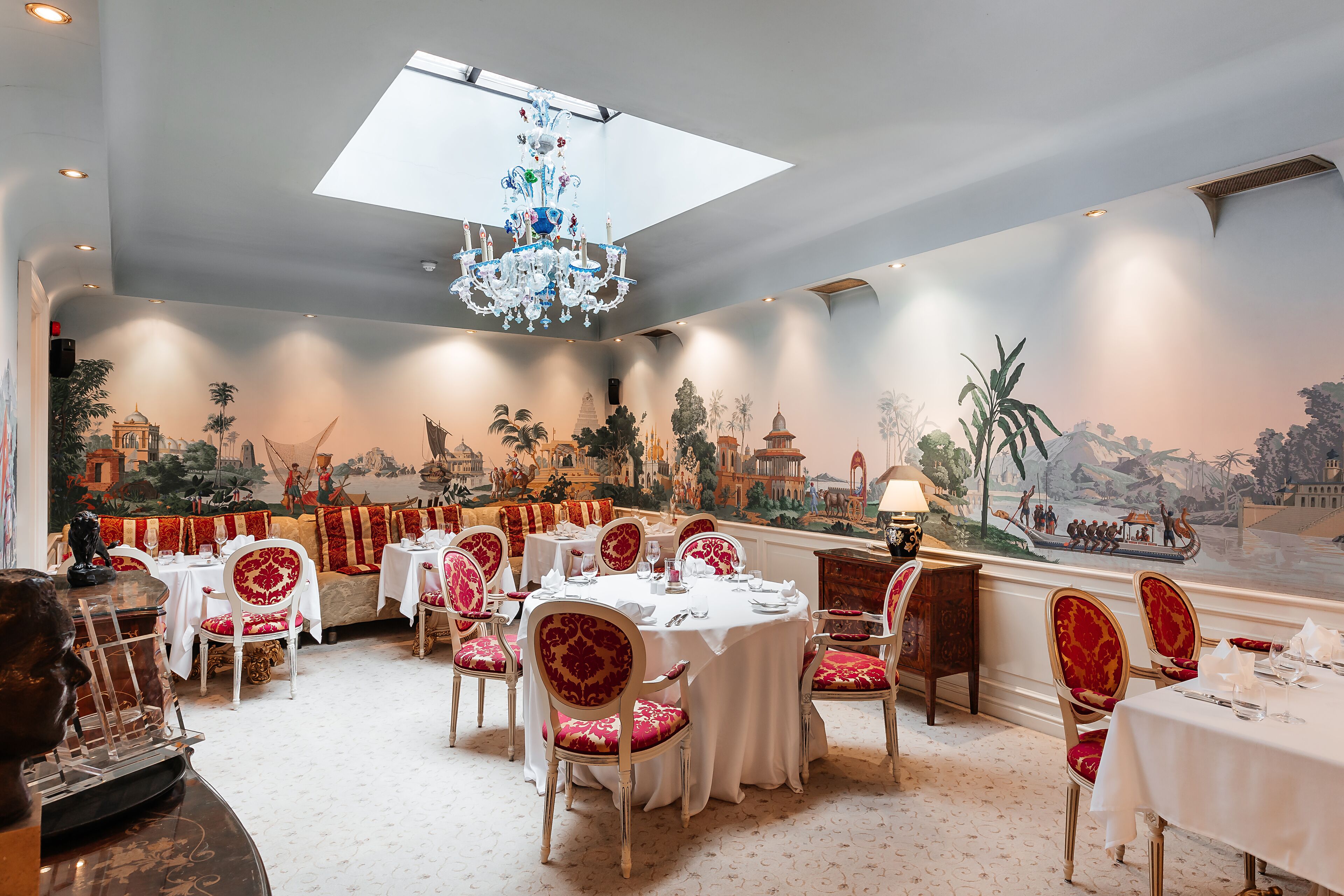 Een restaurant met ronde tafels, rode beklede stoelen en een muurschildering van een stadsgezicht.