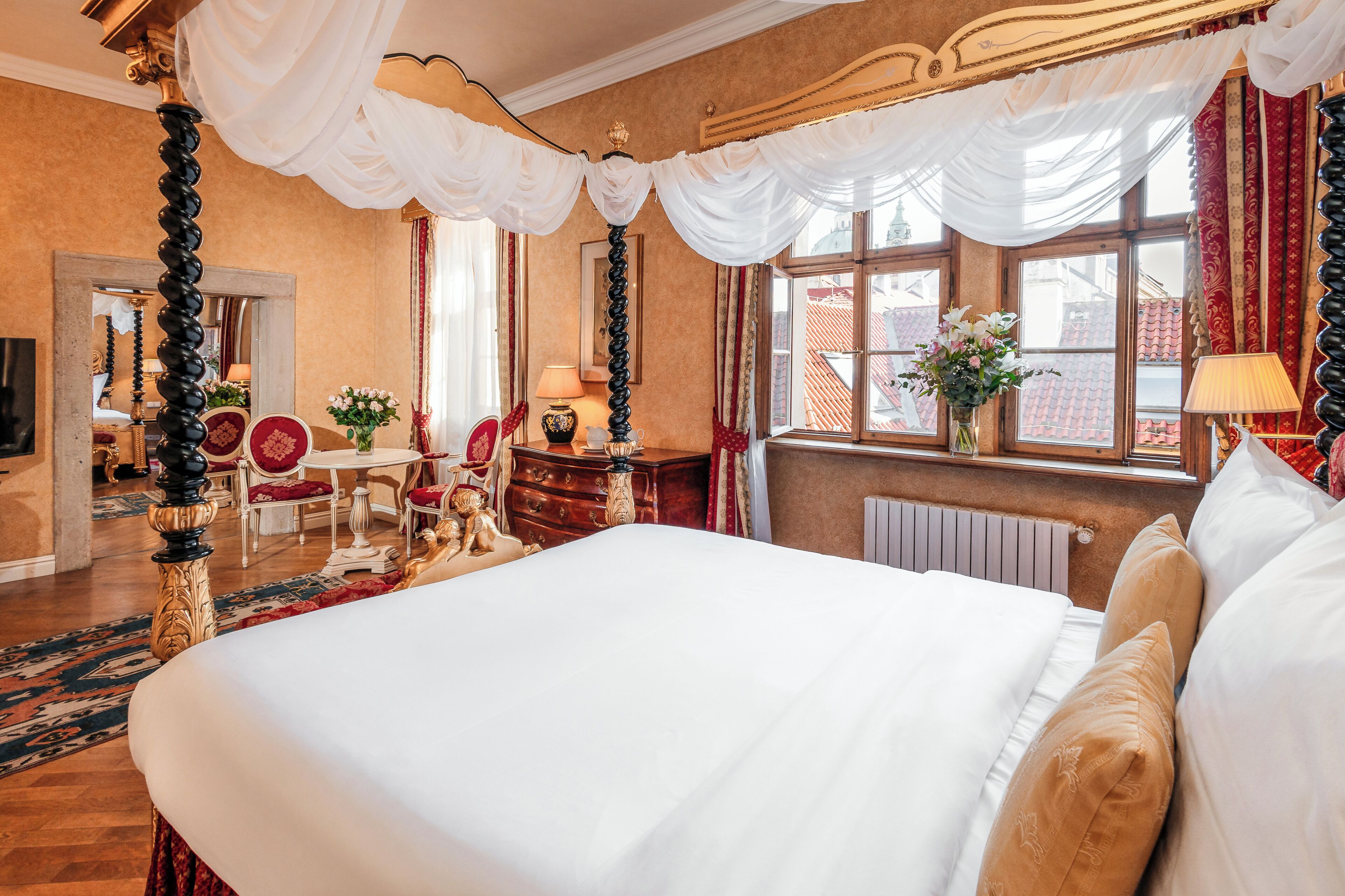 A four-poster bed with white linens, a sitting area with a table and chairs, and a view of the city through large windows.