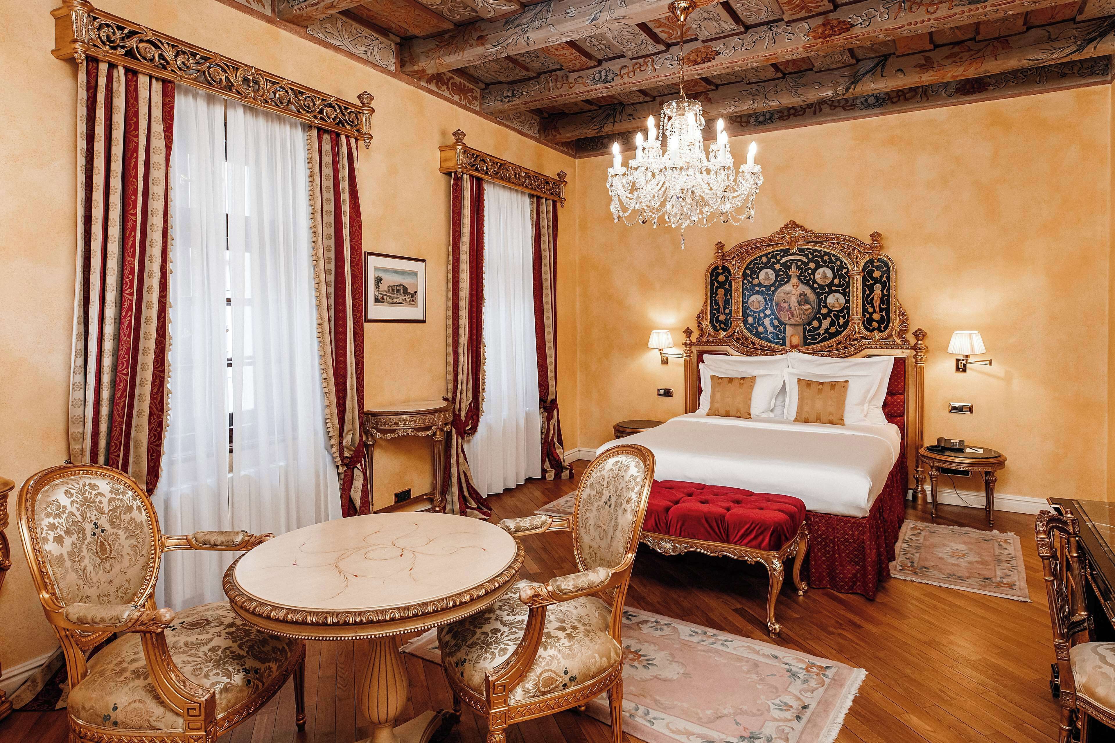 A luxurious bedroom with a large bed, ornate headboard, chandelier, and classic furniture.