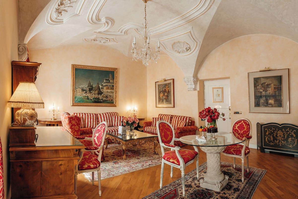 Ein geräumiges Wohnzimmer mit hoher Decke, aufwendigen Deckenverzierungen und einem Kronleuchter. Es gibt mehrere Sitzbereiche, darunter eine Sofagarnitur, Sessel und ein Esstisch.