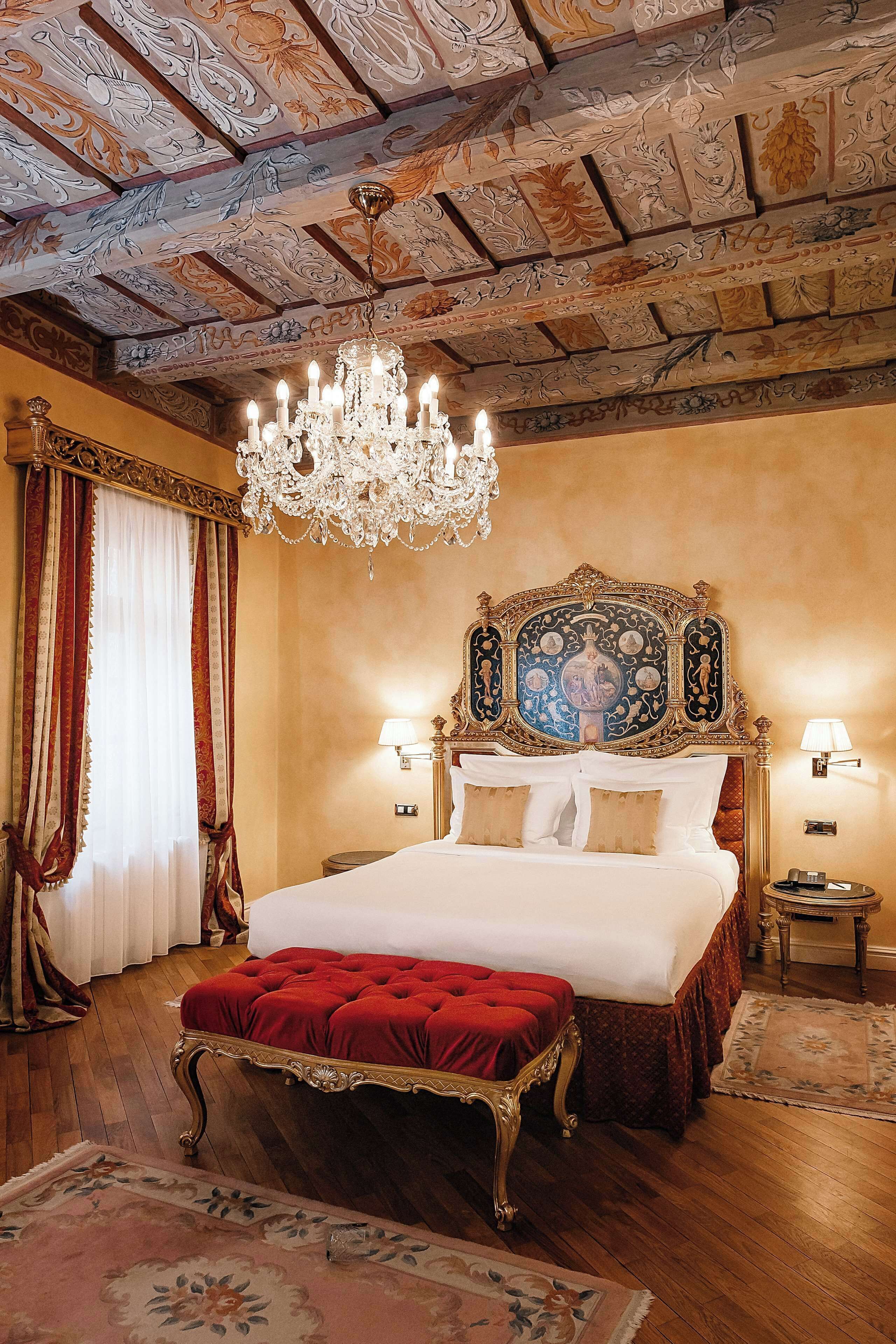 A luxurious bedroom with an ornate ceiling, a large bed with red upholstery, and a chandelier.