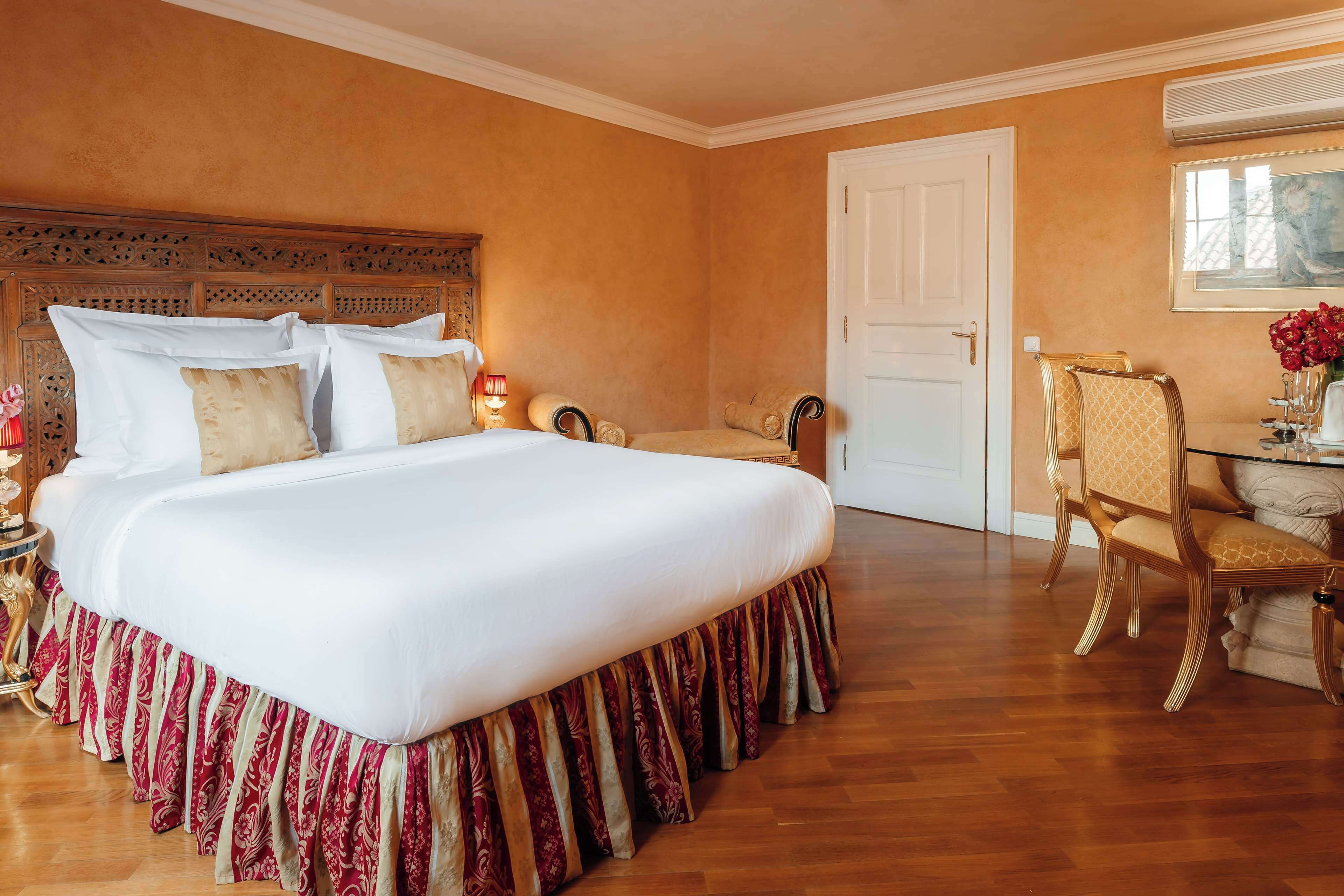 Un dormitorio amplio con una cama grande, un cabecero de madera, una zona de estar con una silla y una mesa con un florero.