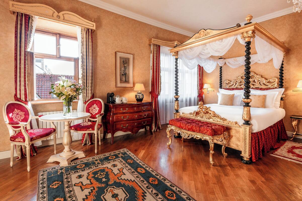 Ein Bett mit Baldachin, ein Esstisch mit Stühlen, eine rot gepolsterte Bank und ein gemustertes Teppich in einem Raum mit Holzböden und einem großen Fenster.