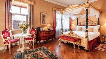 A four-poster bed, a dining table with chairs, a red upholstered bench, and a patterned rug in a room with wooden flooring and a large window.