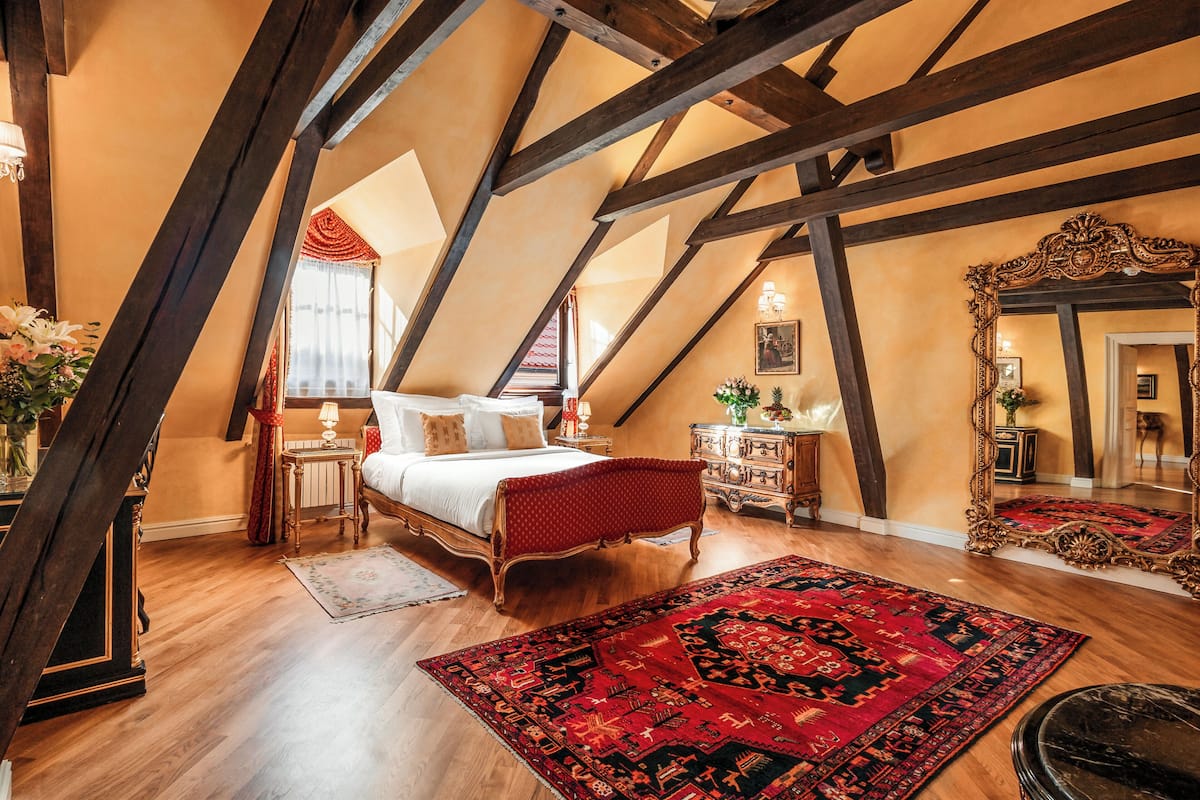 Ein Schlafzimmer mit Holzbalkendecke, einem Bett, einem roten, gepolsterten Kopfteil, einer Holzkommode und einem großen, verzierten Spiegel.