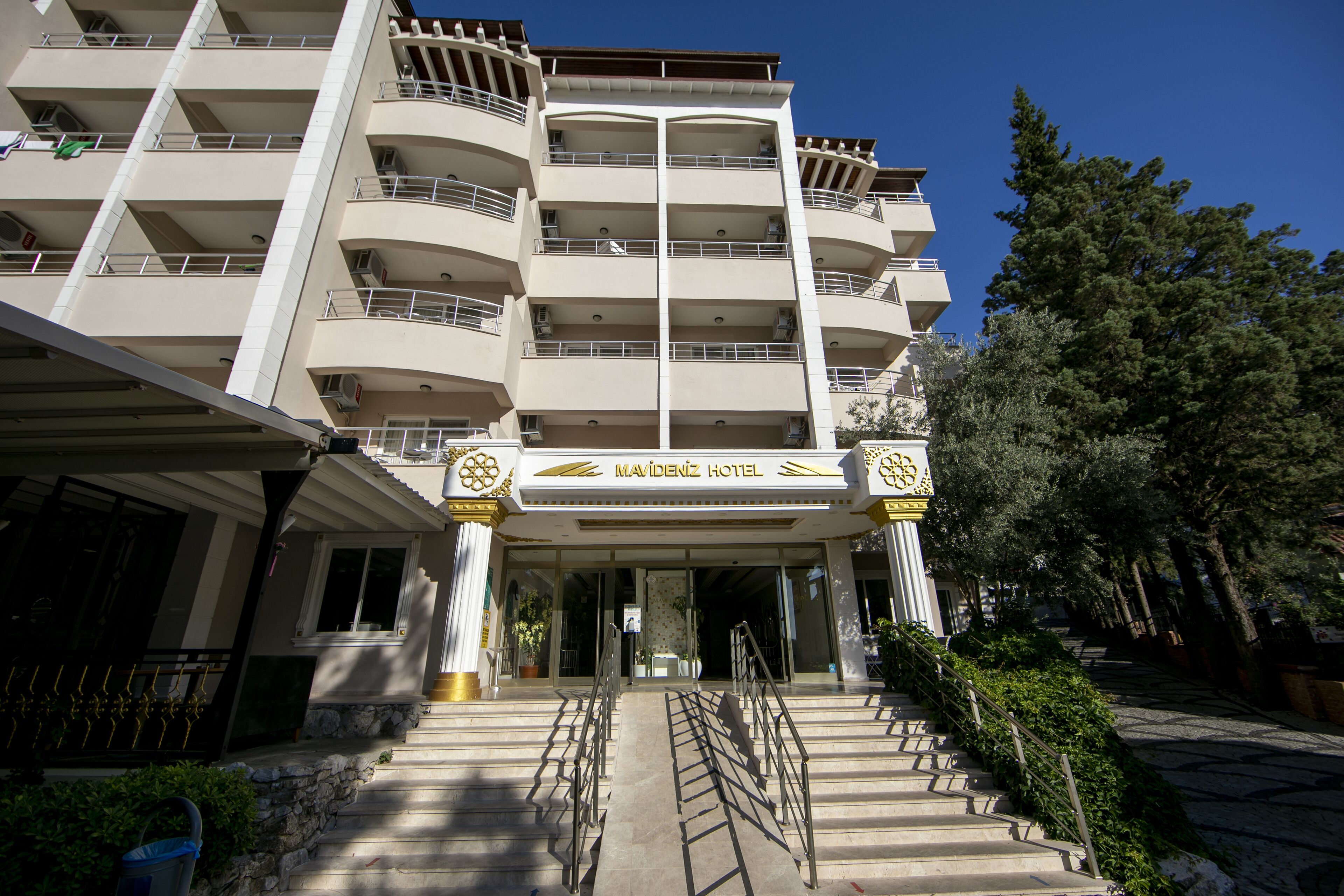 A multi-story hotel with a central entrance, balconies on each floor, and a clear sky.