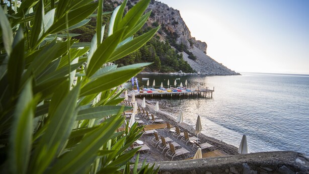 A coastal area with a rocky cliff, a pier with umbrellas and chairs, and a calm sea. - Mavi Deniz Otel - All Inclusive (Marmaris)